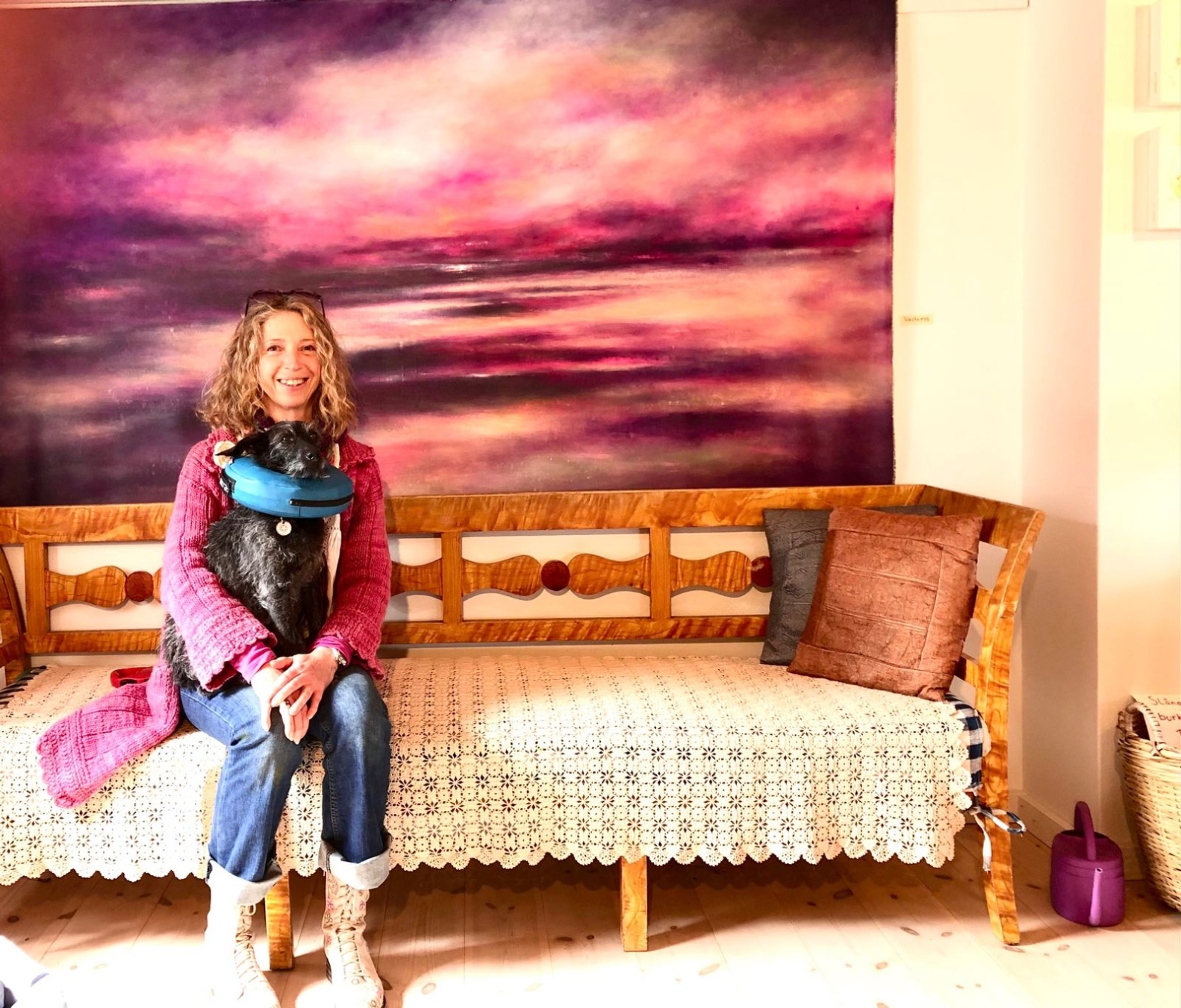 Konstnären Nina Jacucci sitter framför en av sina målningar med hunden Tassa i knät som är nyopererad, därav den blåa uppblåsta kragen.