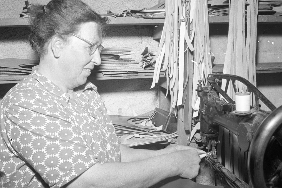 En kvinna sitter och syr någonting i Toffelmakeriet i Åhus. Vad gör hon? Vem är hon? Hur många par tofflor kunde tillverkas på en vecka? Fotografiet är från 1953.