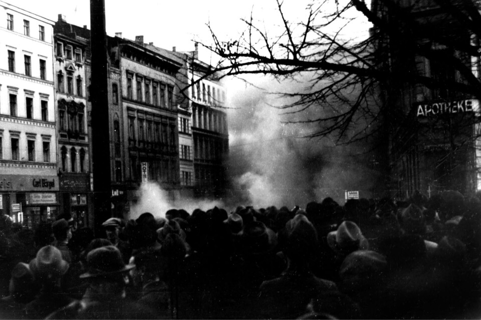 Arkivbild från Kristallnatten i Tyskland den 10 november 1938. Stor folksamling i förgrunden och rök som stiger mot skyn en bit längre fram.