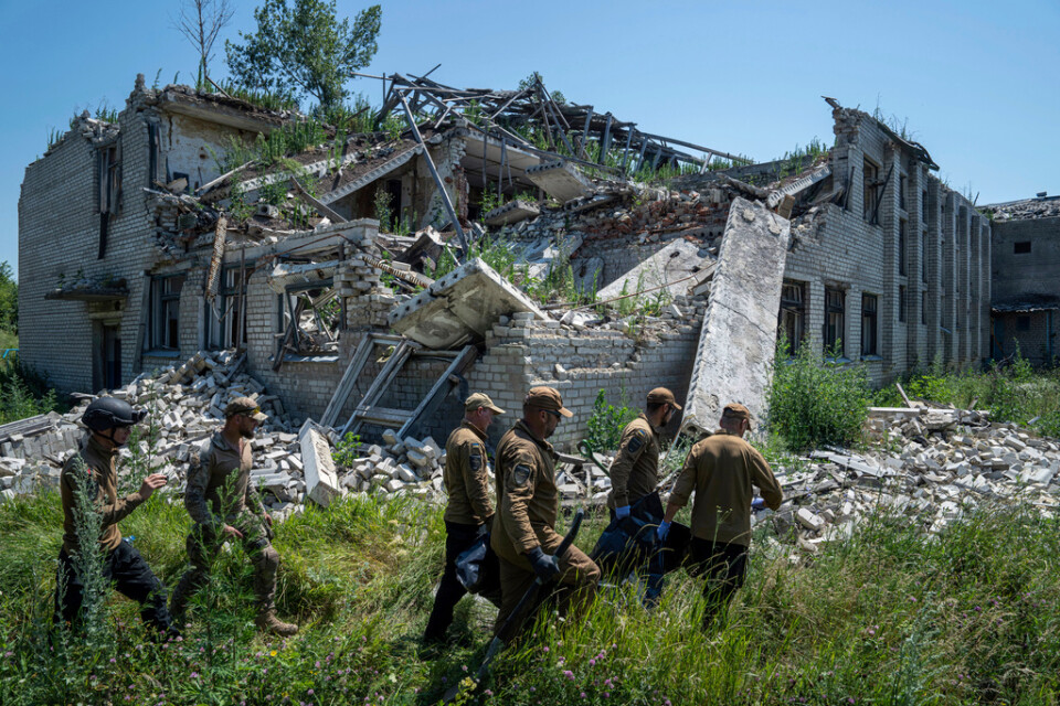 Kvarlevorna av en rysk soldat bärs bort från en krater som han hittades i, invid en förstörd skolbyggnad i orten Virnopillja. Bilden togs den 6 juli. Arkivbild.