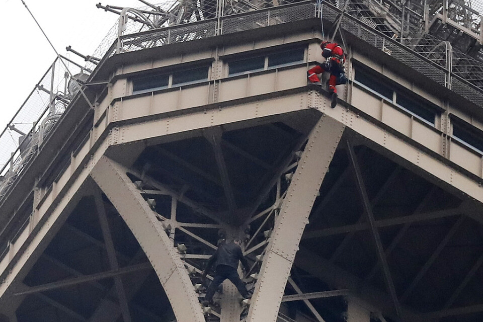 Den svartklädde klättraren syns mellan två balkar i bildens nederkant. Ovan, en rödklädd räddningsarbetare.