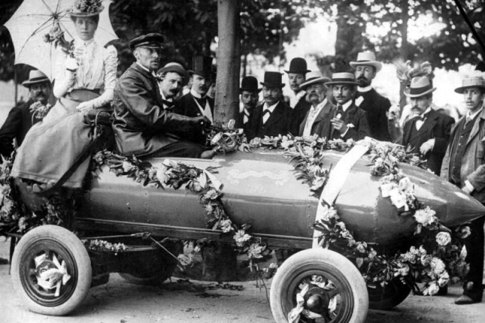 Camille Jenatzy sitter i bilen som kallades "Jamais Contente", det vill säga "aldrig nöjd". År 1899 var den bilen först med att köra i 100 kilometer i timmen.