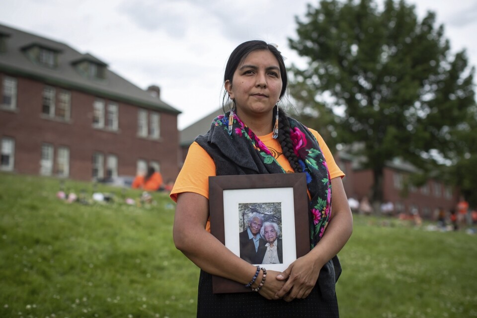Stephanie Gutierrez står utanför den tidigare internatskolan i Kamloops där kvarlevorna hittats, på fotot hon bär syns hennes farmor som tvingades gå i skolan.